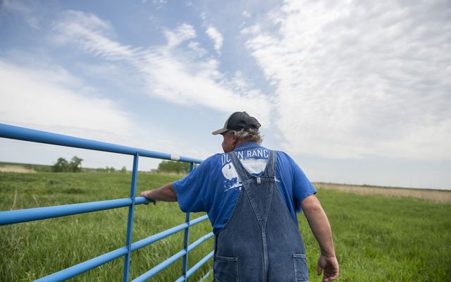 man in overalls and a blue shirt reading "Dunn Ranch" is seen from behind as he moves a stock gate in a green field