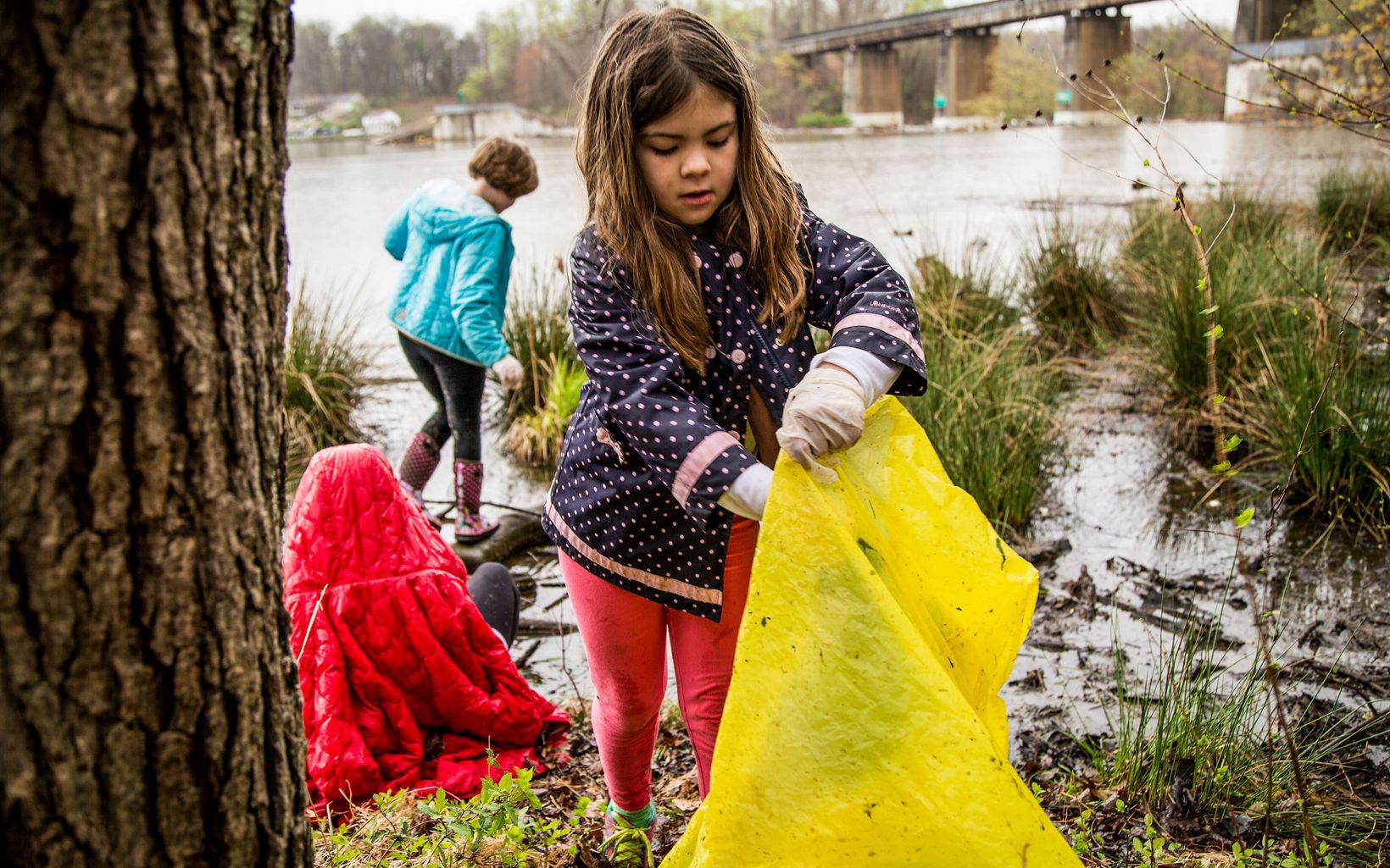 A young girl wearing a navy blue and pink polka dotted rain coat puts trash in a large yellow bag.