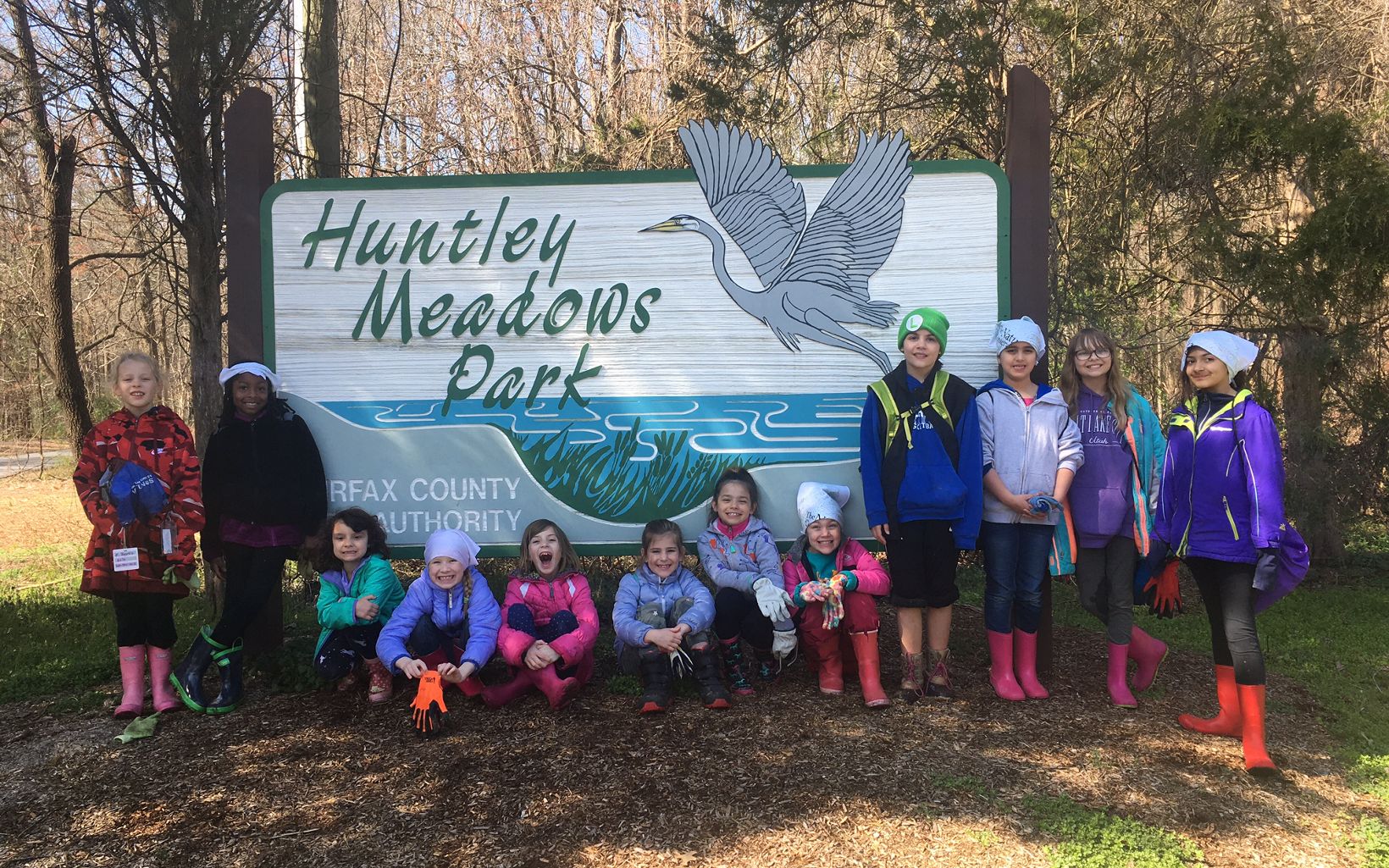 A smiling group of Brownie Scouts. Twelve girls lined up in front of a large sign reading "Huntley Meadows Park."
