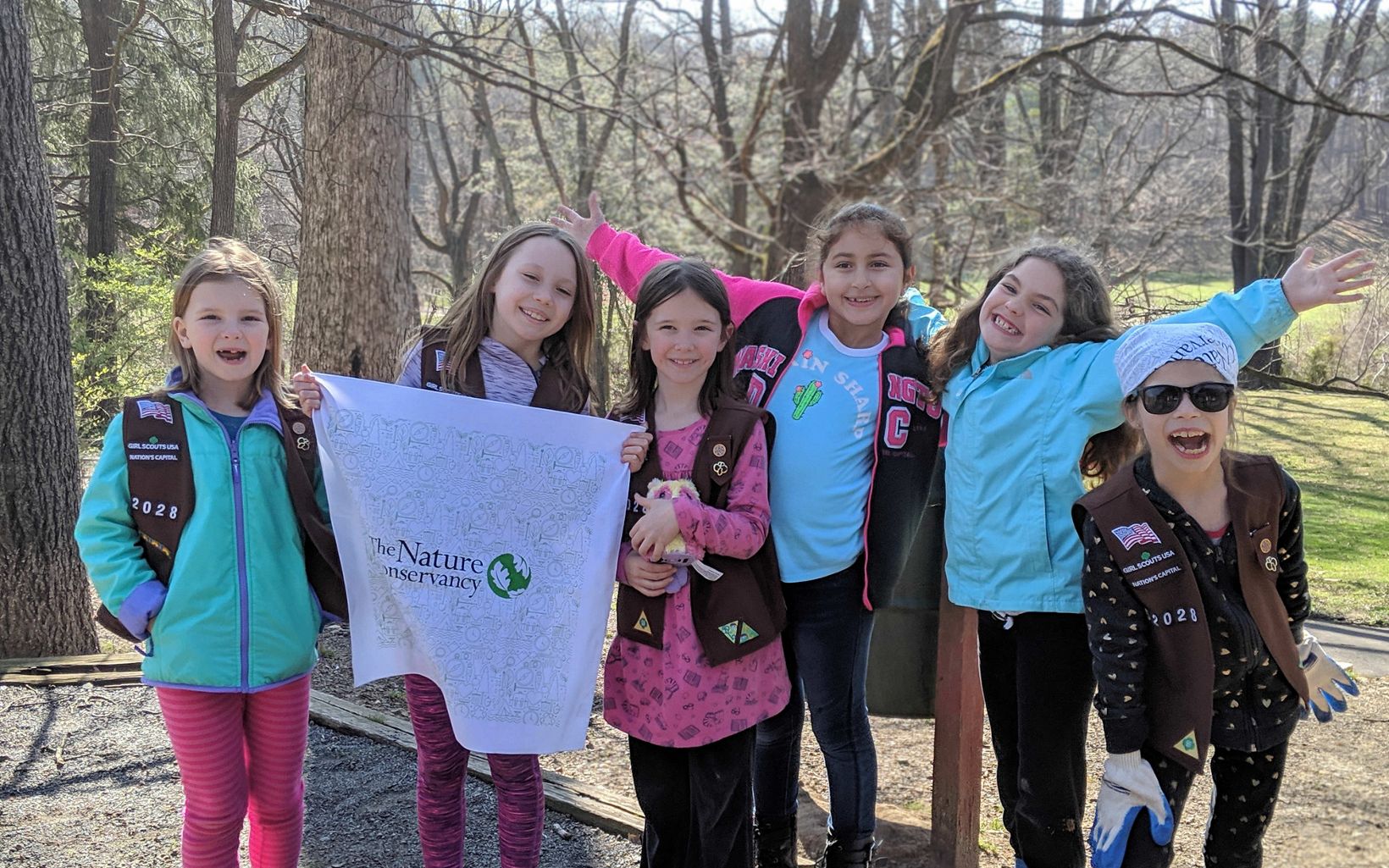 A smiling group of Brownie scouts. Six girls standing together in front of a grove of trees.