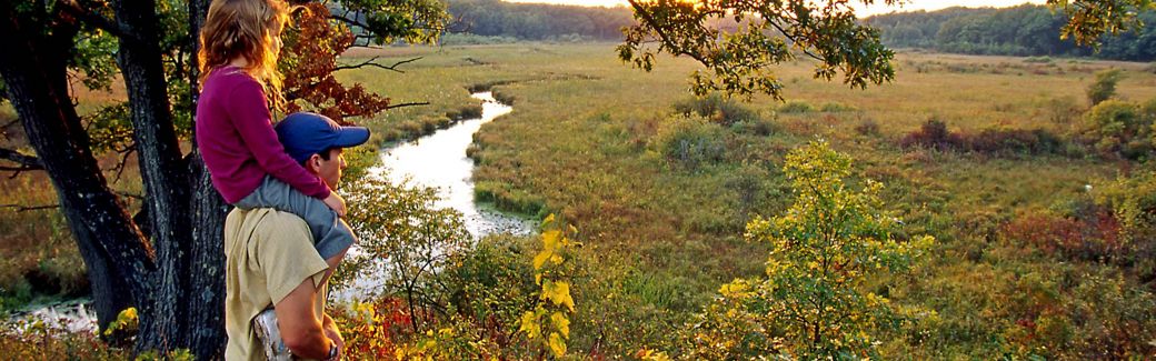 Father with daughter on shoulders looks across wetlands and winding river to forests beyond as setting sun makes fall colors of vegetation glow.