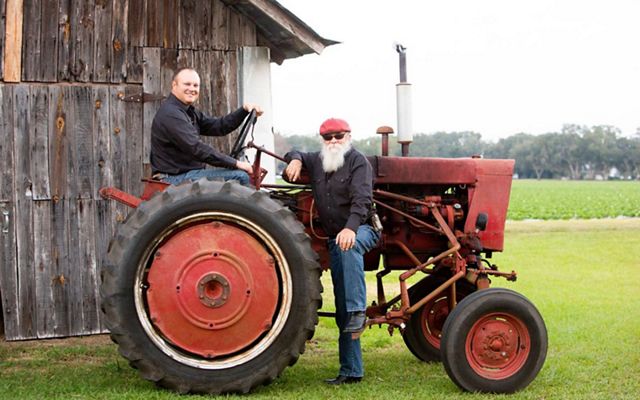 A smiling farmer sits on a red, vintage tractor in front of an old barn, while another farmer with a big white beard  stands next to the tractor leaning his arm on it.