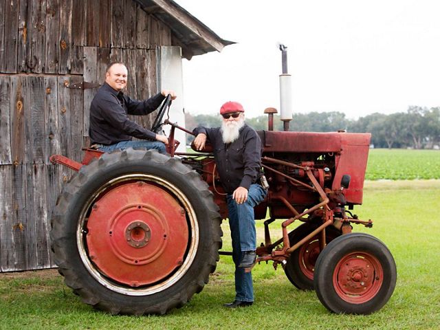 A smiling farmer sits on a red, vintage tractor in front of an old barn, while another farmer with a big white beard stands next to the tractor leaning his arm on it.