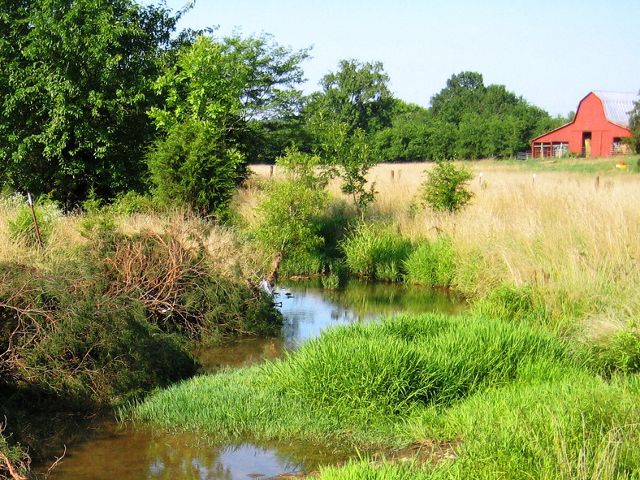 A stream winds through a field, away from a small red barn.
