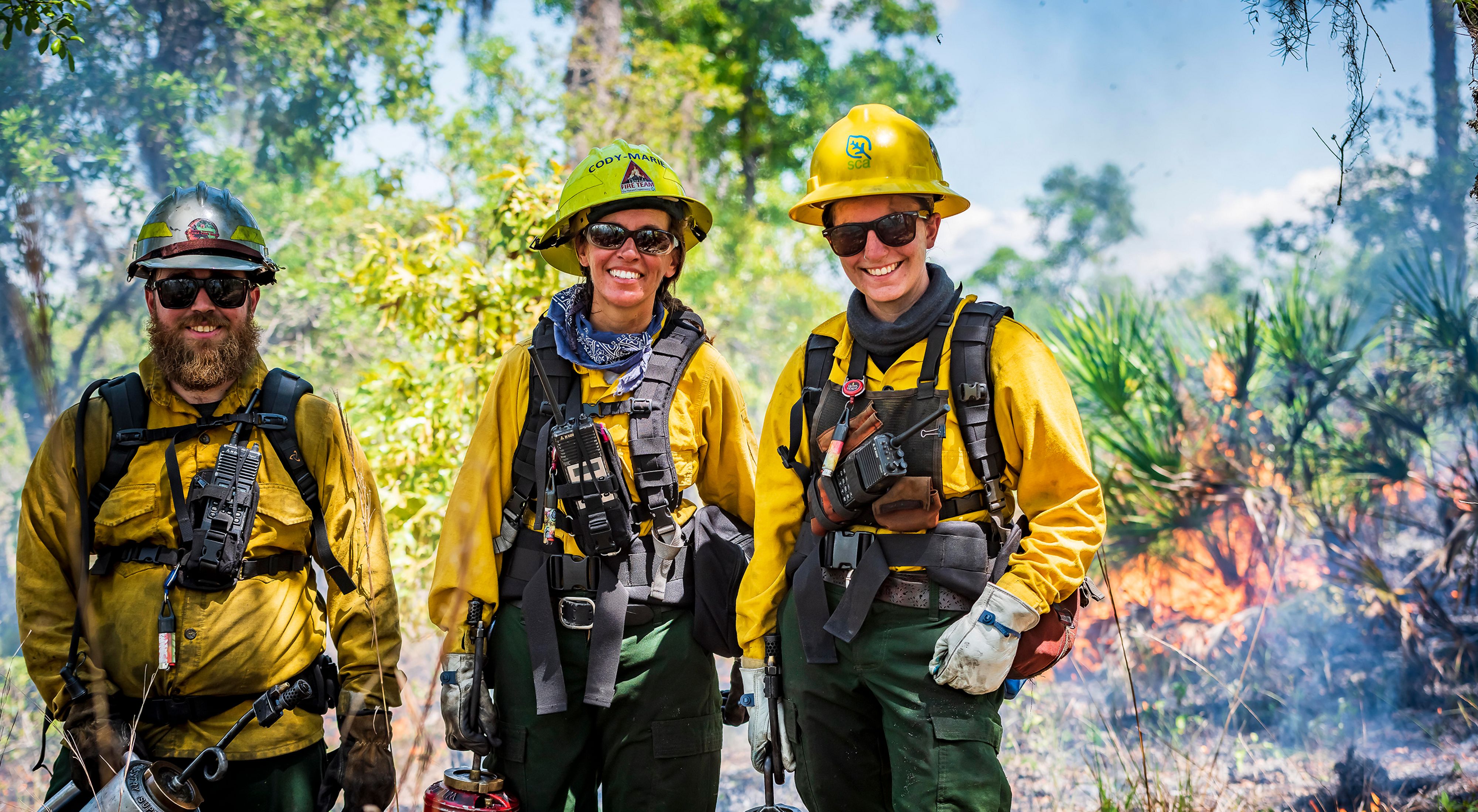 Three people in protective fire gear smile at the camera while a controlled fire burns behind them