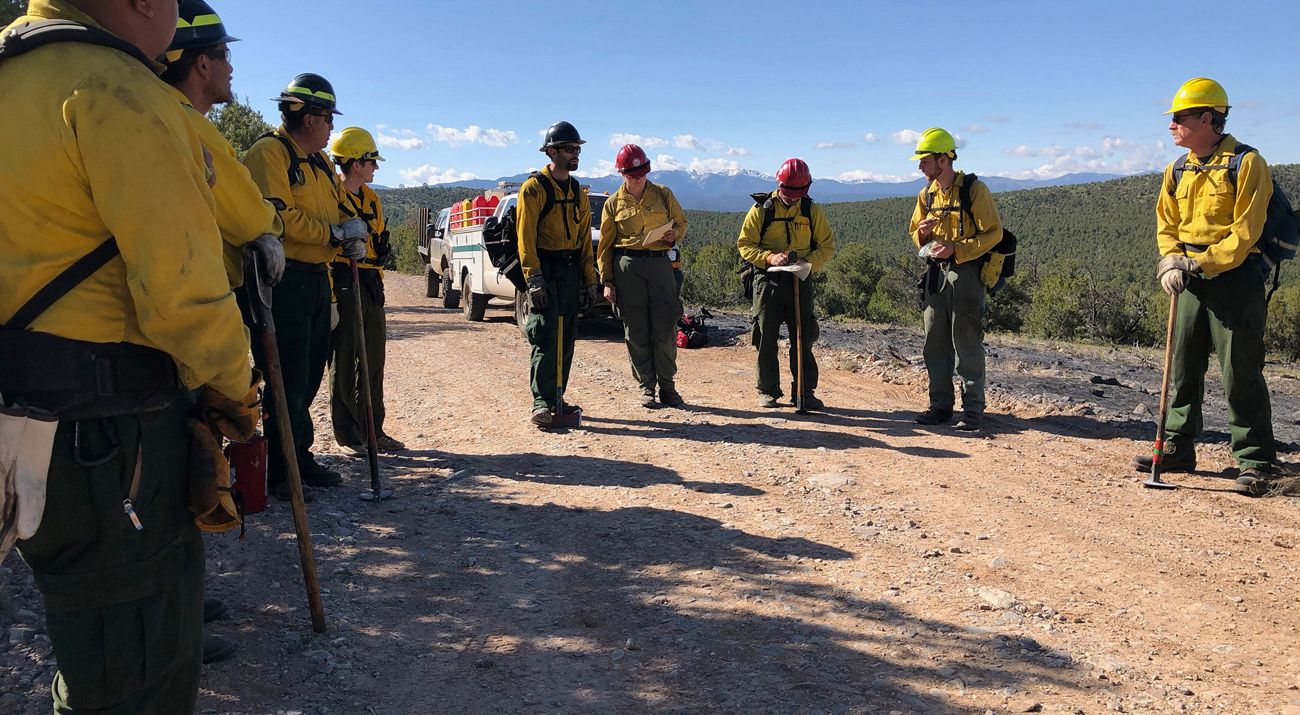 Several people in yellow prescribed fire gear and hardhats discuss plans.