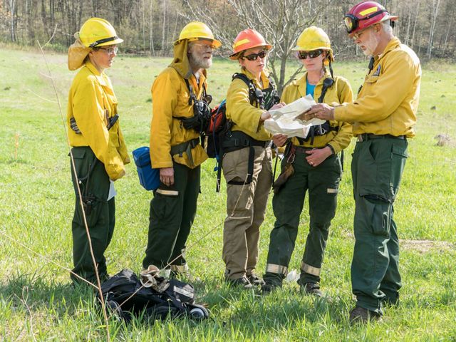 Five people dressed in yellow Nomex suits, helmets, goggles and other fire gear consult maps while working on a prescribed fire in Wisconsin.