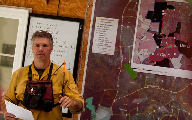 A man stands in front of a wall with maps tacked to it, leading a briefing before a controlled burn.