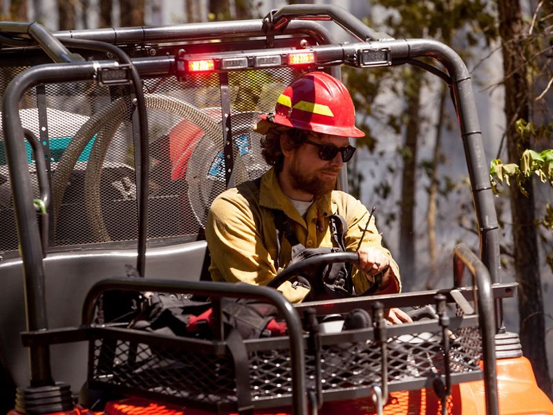 A man wearing a yellow shirt and red hard hat drives a small fire truck. The open sided all terrain vehicle contains hose and water tanks.