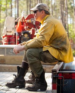 A man wearing sunglasses and a soot streaked yellow shirt sits and rests at the end of a controlled burn.