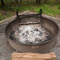 Ash covers the bottom of a large, round metal fire pit. A large piece of firewood lays on the ground in front of the fire pit.