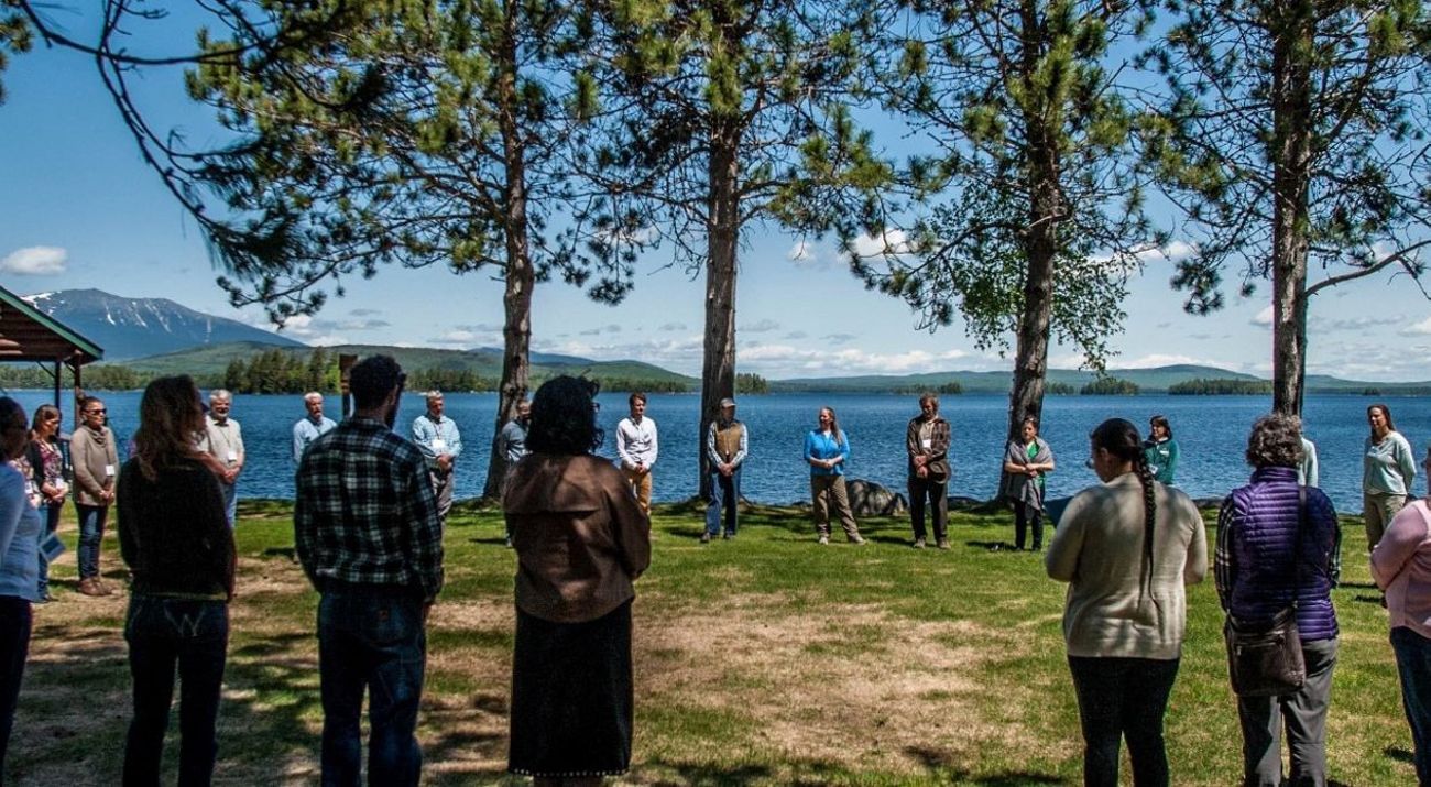 A group stands in a circle with a lake behind them.
