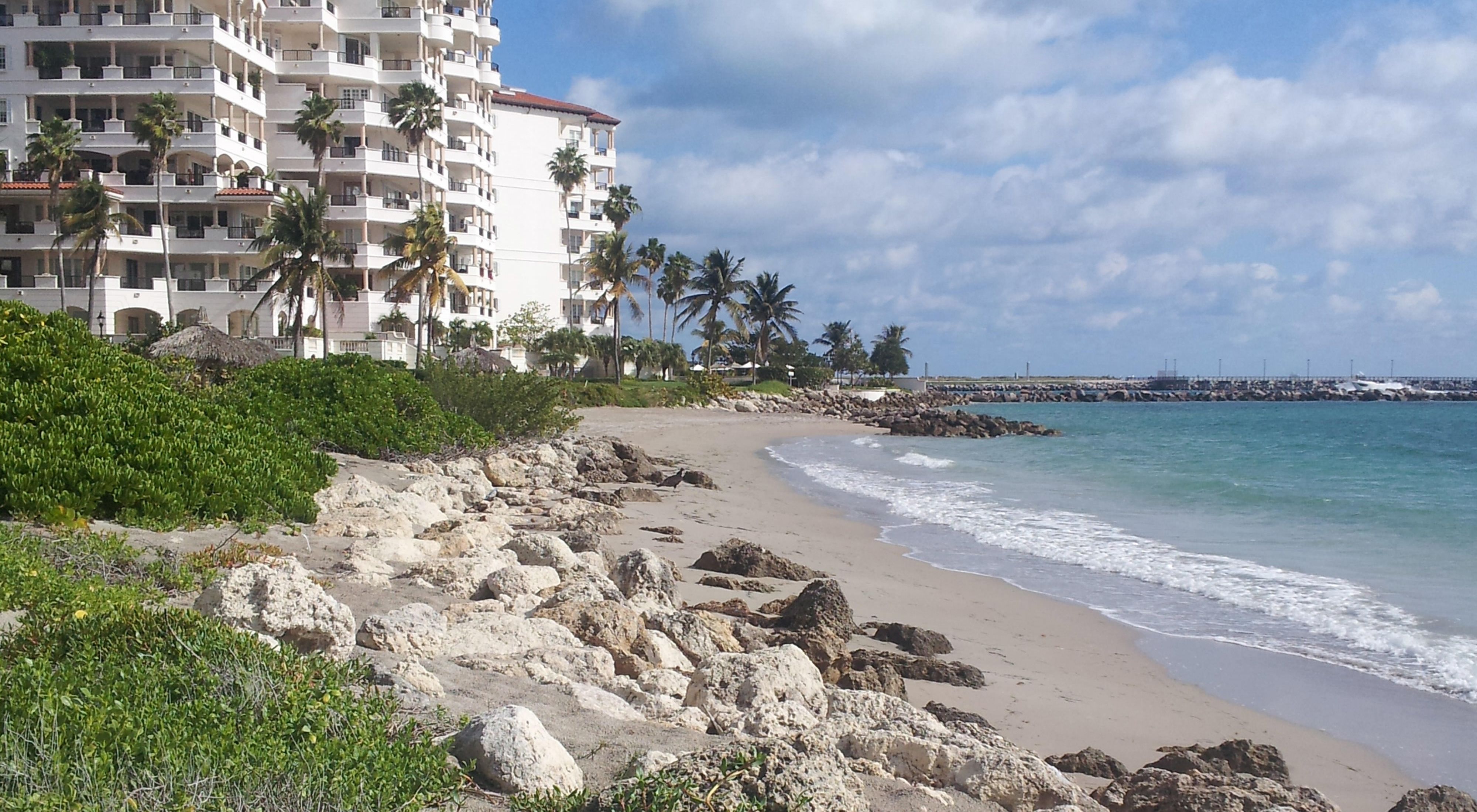 A highrise condominium building sits very near to the ocean shoreline with blue water, rocks and a sandy beach in Fisher Island, located in the Miami, Florida metropolitan area.