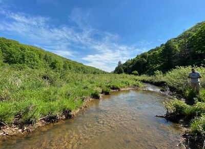 A man stands at the edge of a wide, shallow stream. Two mountain ridges meet in a vee in the background framing bright blue sky.