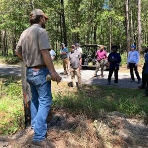 A person talks to a gathering of people near a forest.