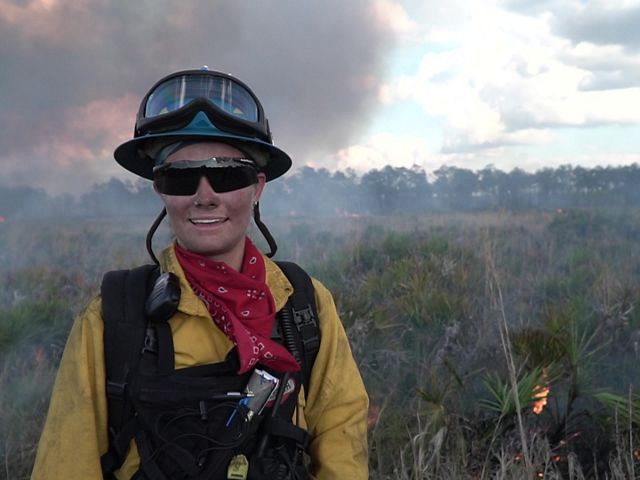 Amanda Lindsay in fire gear with fire and smoke in the background.