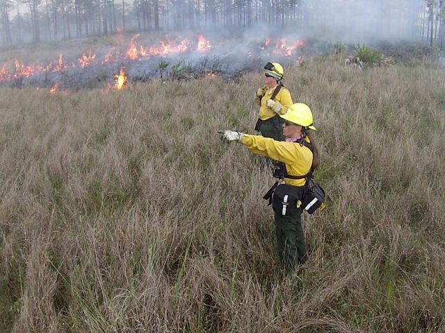 Two women stand in a grassy field wearing fire gear with flames in the background.