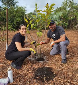 Two people smile at the camera, their gloved hands holding a young, newly planted tree.