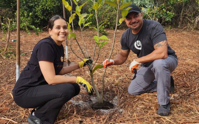 Two people smile at the camera, their gloved hands holding a young, newly planted tree.
