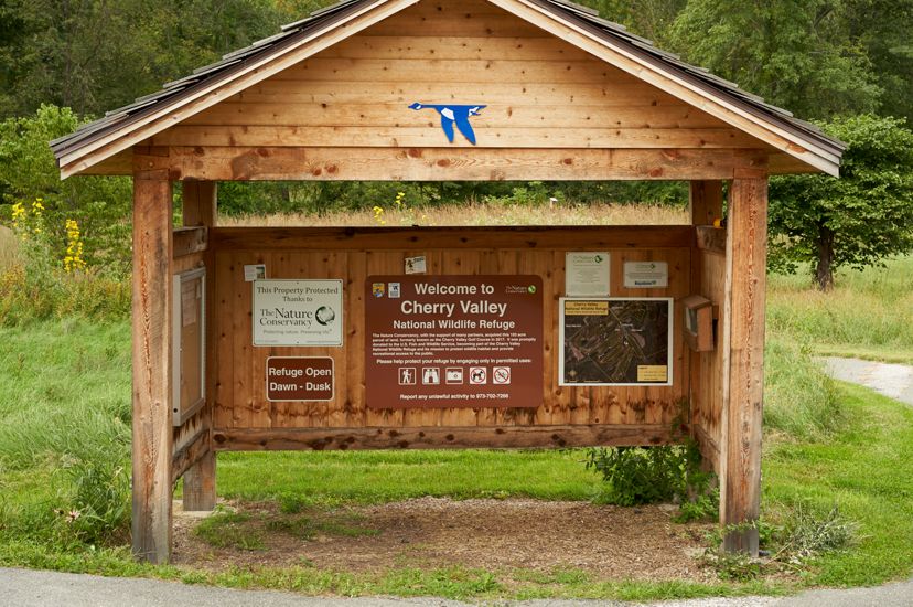 A triangular shaped wooden structure stands at the entrance of a path into the forest. The structure contains various signage and papers with information.