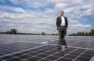 A man stands in the middle of an array of solar panels under a bright blue sky filled with white puffy clouds.