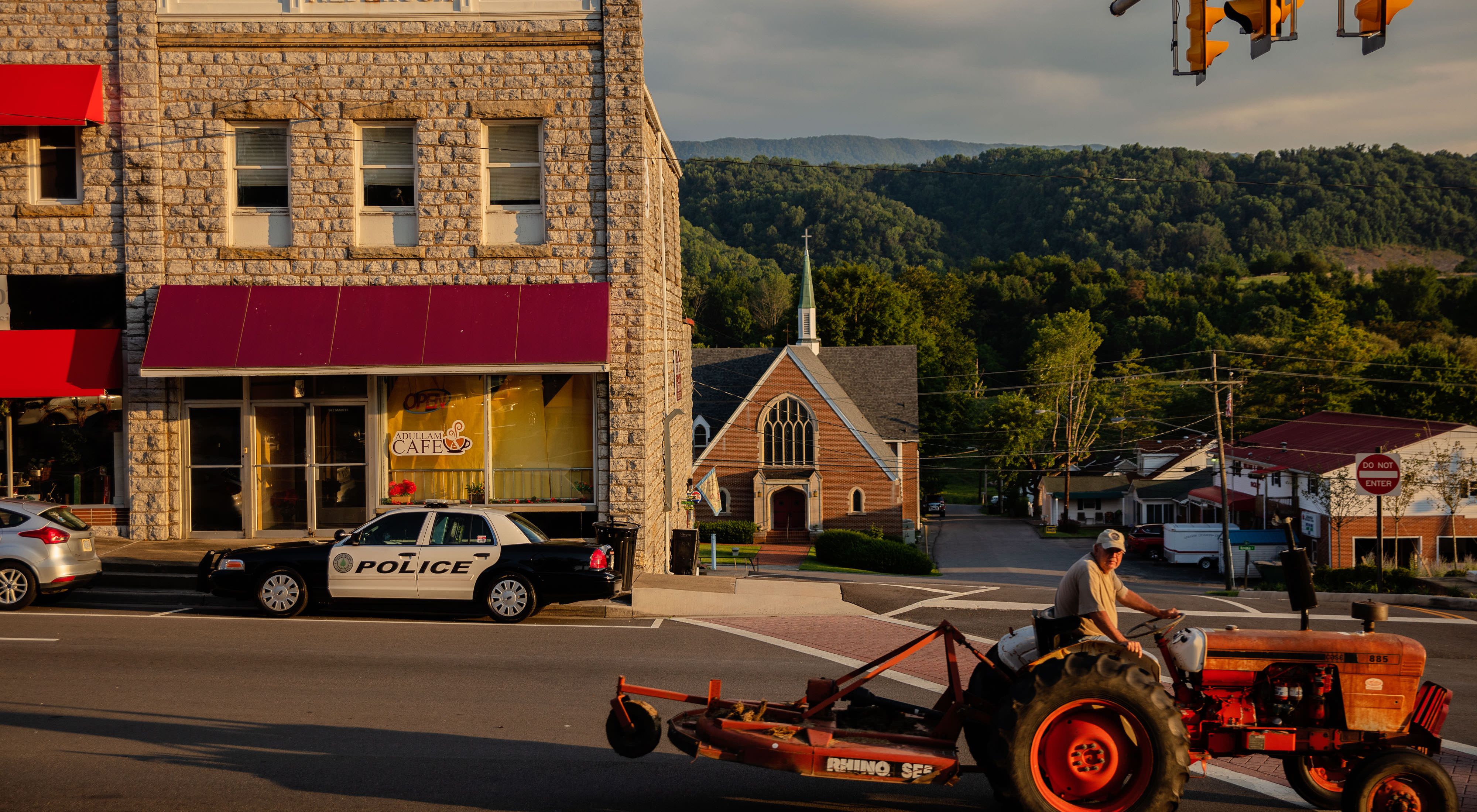 Street scene in St. Paul, VA. A red tractor drives past stone clad storefront. A police car is parked on the street. A church and other buildings are visible with a tall mountain in the background.