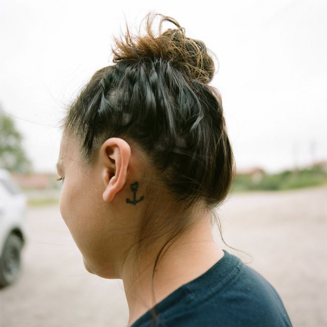 Woman with dark, wispy hair pulled into a bun, tattoo of an anchor shown behind her ear