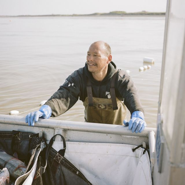 Man stands in brown water in waders, pushes boat towards camera, buoys atop a net extend in the background