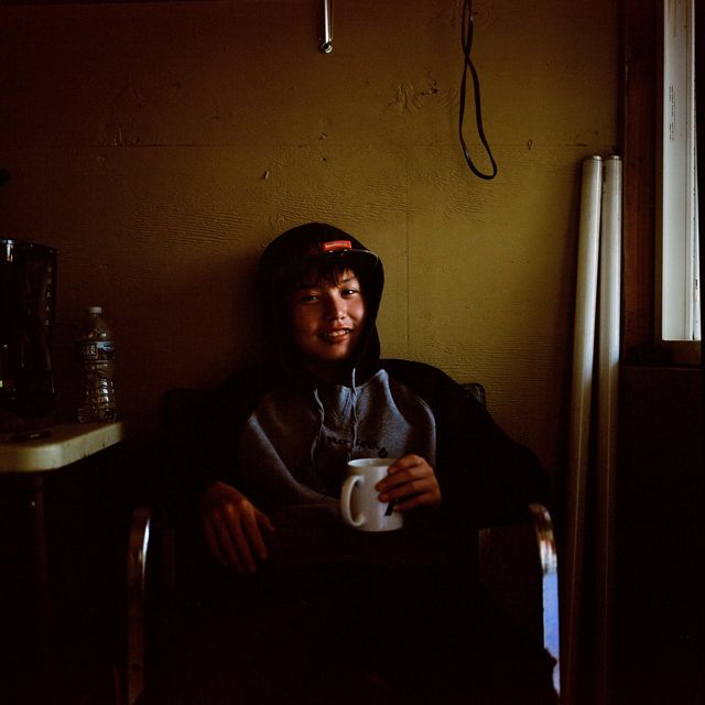young boy in hoodie sits indoors by window with mug, looks at camera