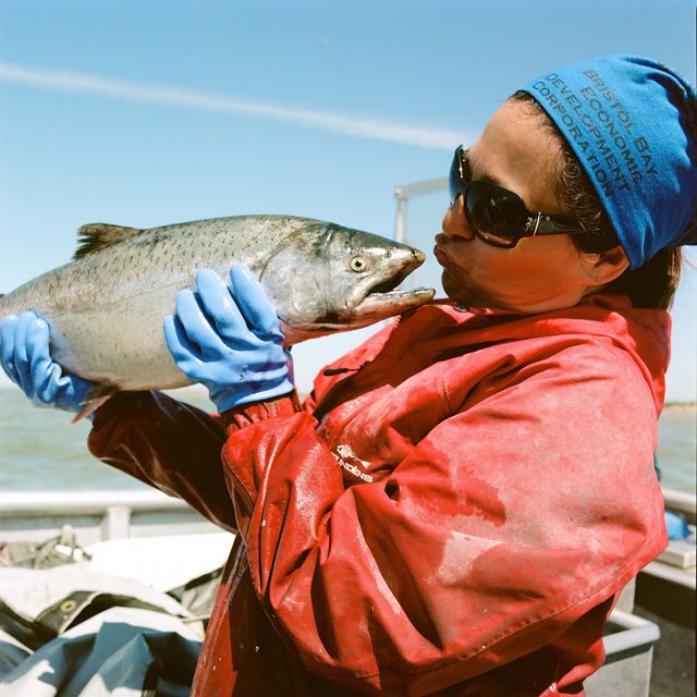 Fisher in red jacket holds silver-colored fish up to her face and almost kisses it. In boat on water