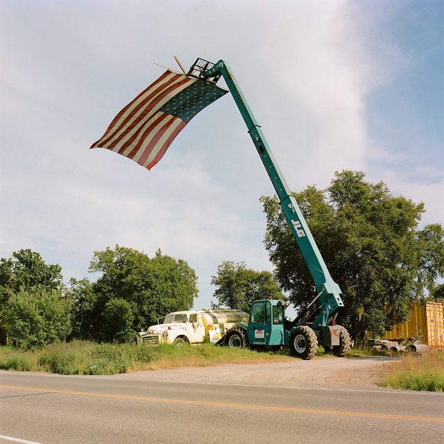 by the side of a road, a crane holds an American flag vertically that blows in the window below the top of the crane