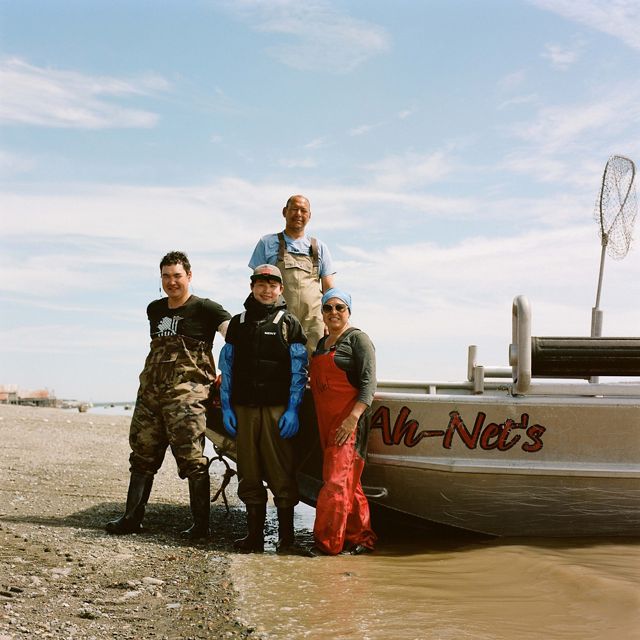 4 people in waders stand beside boat pulled onto rocky shore, smiling for picture