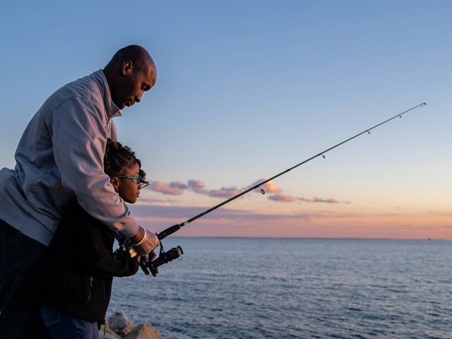 Un padre está parado detrás de su hija y la ayuda a manejar una caña de pescar en la costa de Bridgeport, Connecticut.