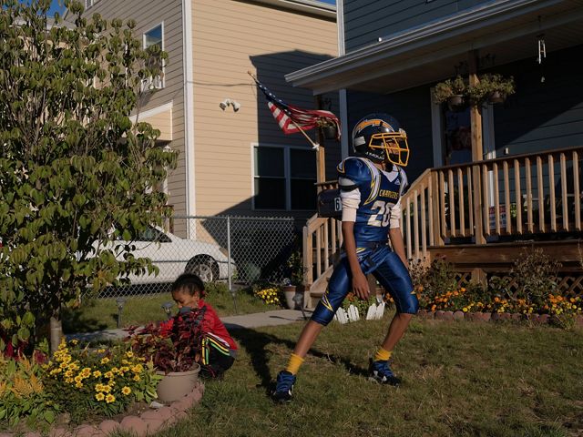 A front yard in Bridgeport, Connecticut shows a young boy near a newly planted tree and his older brother standing nearby in football gear. 