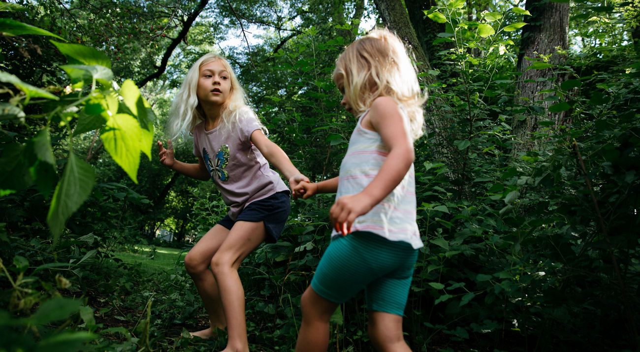 A barefoot girl reaches for the hand of a younger also barefoot girl to draw her through a gap in some bushes.