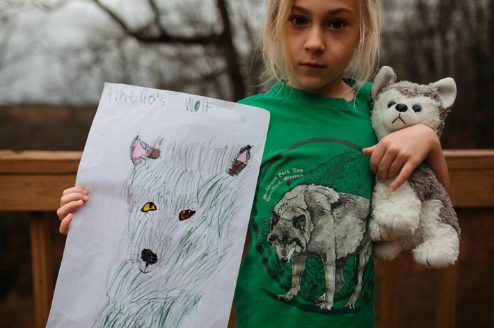 A girl holds a stuffed wolf in one arm and holds up her drawing of a wolf in the other.