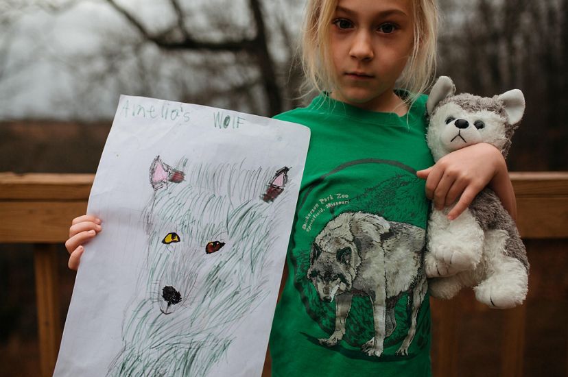 A girl holds a stuffed wolf in one arm and holds up her drawing of a wolf in the other. 