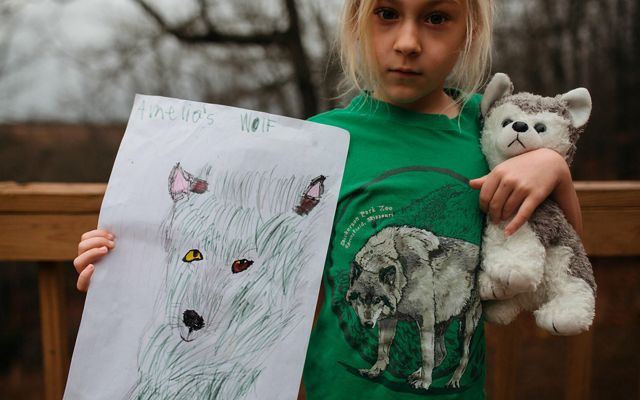 A girl holds a stuffed wolf in one arm and holds up her drawing of a wolf in the other. 