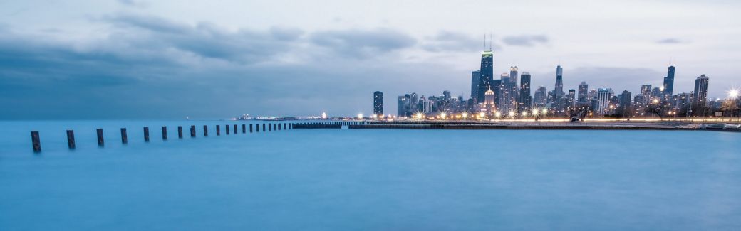 The skyline and lights of the city of Chicago are viewed at twilight with Lake Michigan in the foreground and thick clouds out over the lake.
