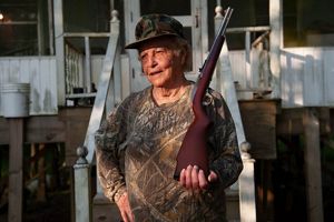 A woman in hunting clothes poses with a rifle.