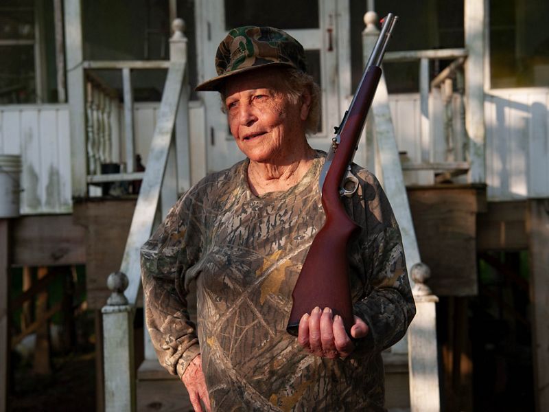 A woman in hunting clothes poses with a rifle.
