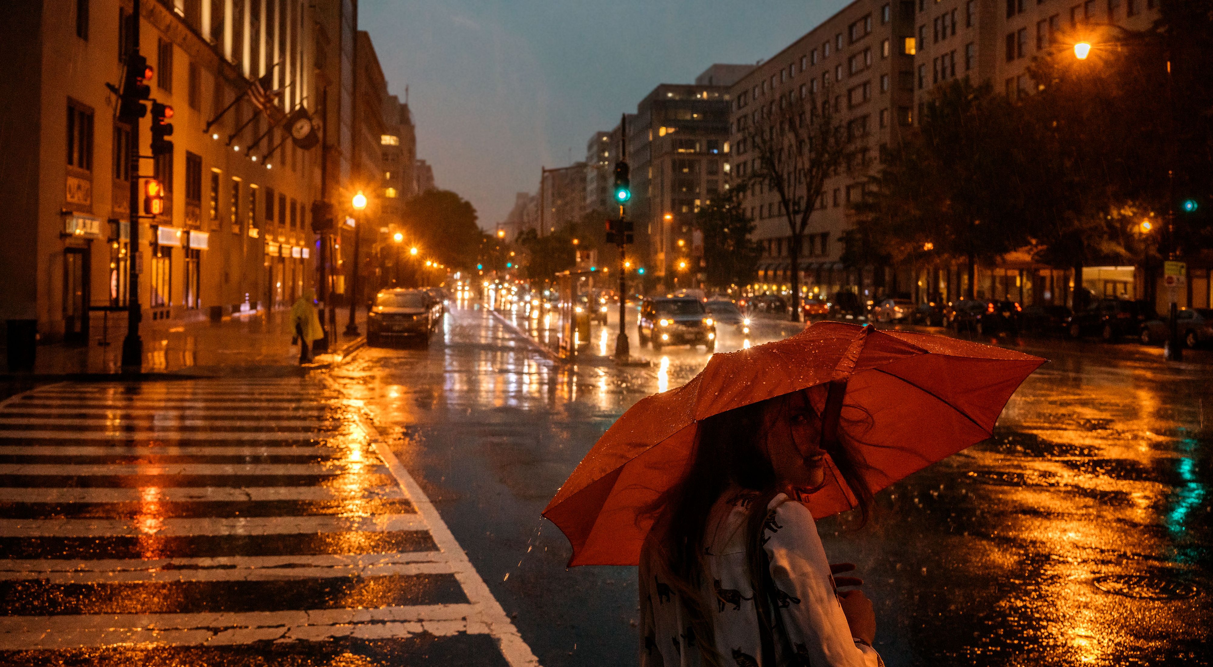 A person with a red umbrella crosses a city street at night.