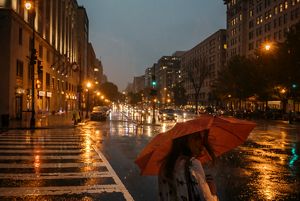 A woman holding a red umbrella waits to cross the street. Street lights and headlights are reflected on the wet pavement.