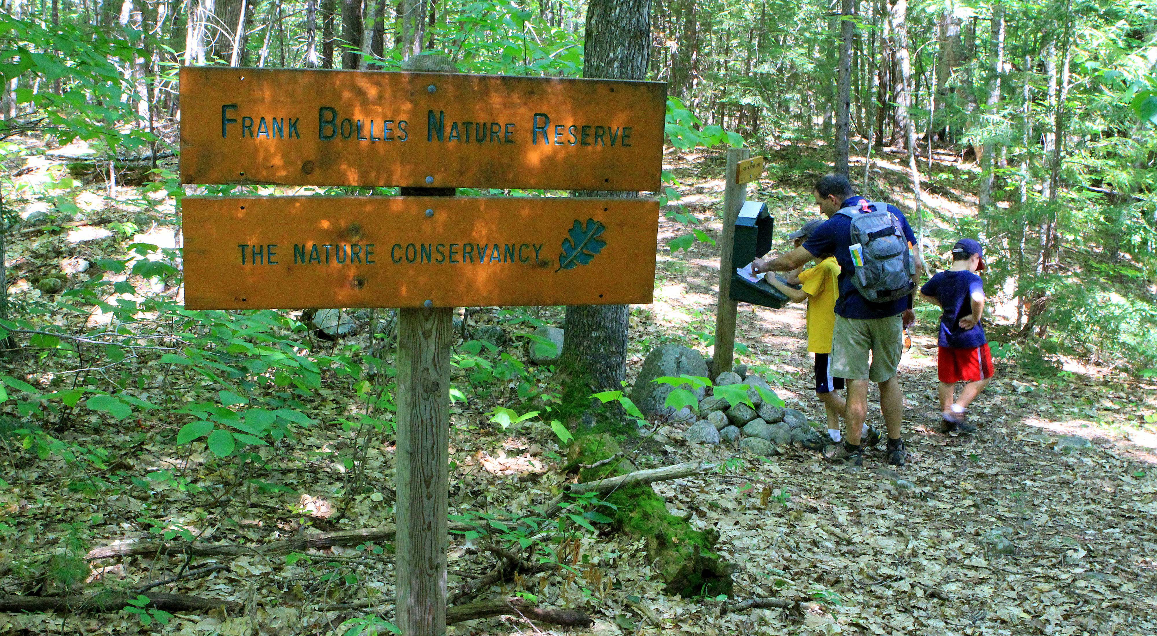 A man and two boys look at a sign-in sheet at a register box in the woods. A wooden sign in the foreground reads Frank Bolles Nature Reserve. The Nature Conservancy.