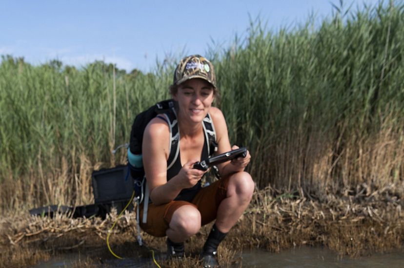 A woman crouches in the shallow water at the edge of a wetland. She holds a joystick pad in her hands, using it to deploy a small rectangular drone through the murky brown water.