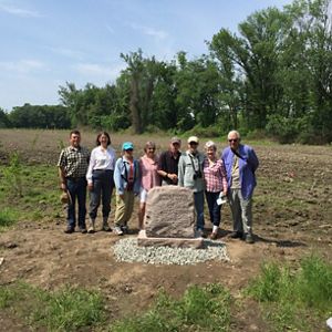A group of people stand in a line smiling around a rock monument in a dirt field with forest in the background.