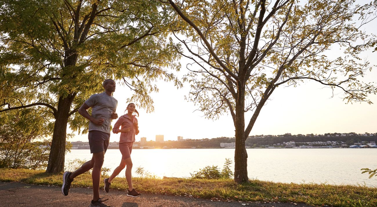 Two people jog along a large body of water under the shade of trees along its banks.