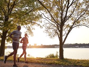 Two people jog along a large body of water under the shade of trees along its banks.