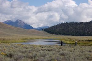 Two people look through binoculars near a river in the mountains.