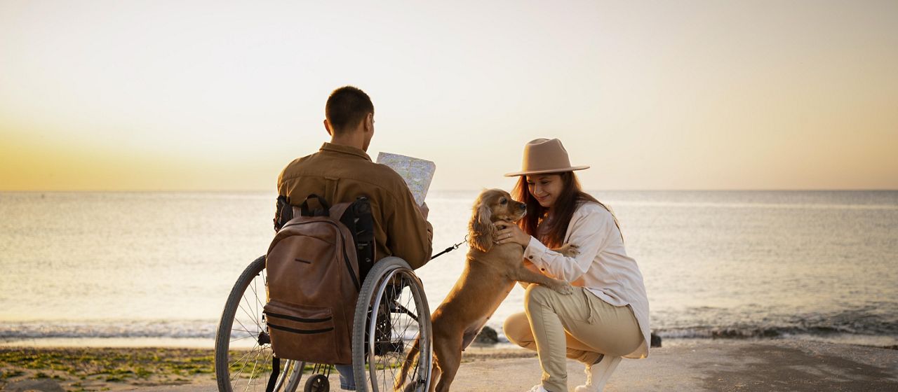 A family enjoys the beach during the sunset. There is a man in a wheelchair and a little dog playing with a woman.